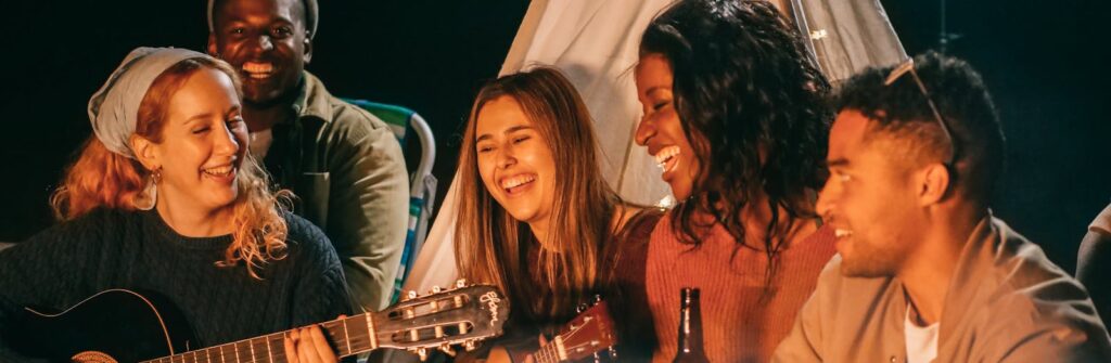 A diverse group of friends playing guitar and ukulele by a campfire at night, having fun and socializing.