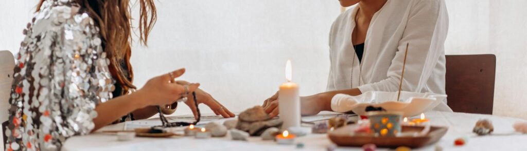 Two women participating in a spiritual ritual with candles and crystals on a table indoors.