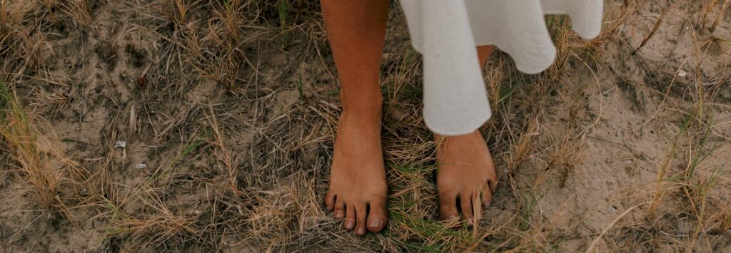 A woman dressed in a white garment stands barefoot on dry, sandy grass, creating a whimsical outdoor scene.