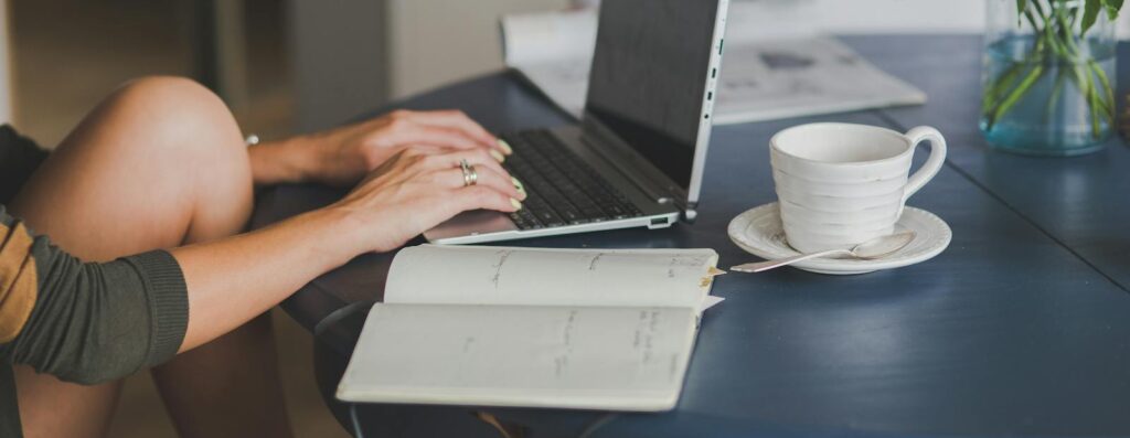 Female freelancer using laptop with coffee at home office desk, surrounded by roses and a planner.