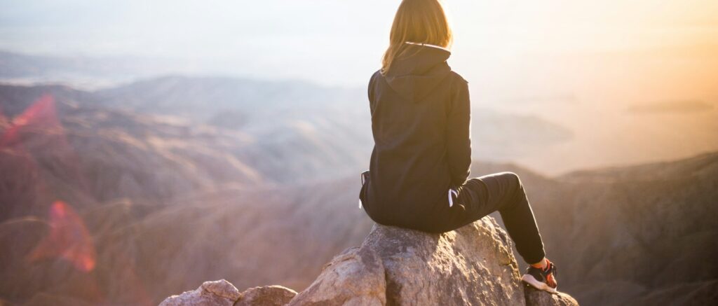 person sitting on top of gray rock overlooking mountain during daytime