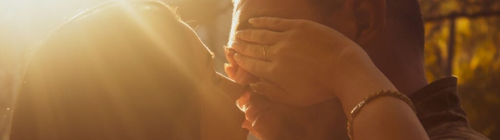 A couple shares an intimate kiss in warm, golden sunlight outdoors, evoking love and romance.